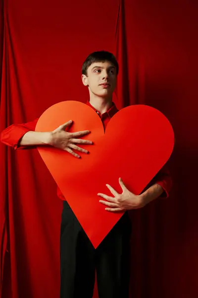 Hombre joven alto en pantalones cortos negros abrazando gran recorte de corazón sobre fondo rojo, día de San Valentín - foto de stock