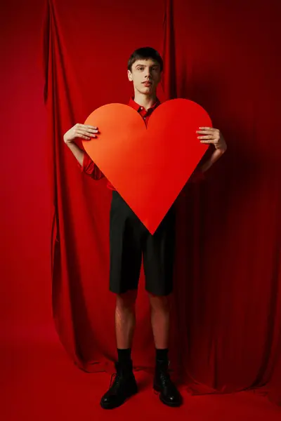 Comprimento total do homem alto abraçando grande caixa em forma de coração no fundo vermelho, dia dos namorados — Fotografia de Stock