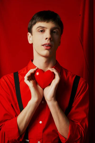 Junger Mann hält kleines Herz zärtlich in den Händen und blickt in die Kamera auf rotem Hintergrund — Stockfoto