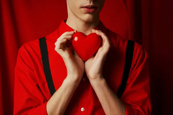 Vista recortada de un joven sosteniendo un pequeño corazón en sus manos sobre un fondo rojo, concepto del día de San Valentín - foto de stock