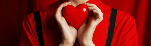 Vista recortada de un joven sosteniendo un pequeño corazón en sus manos sobre un fondo rojo, bandera del día de San Valentín - foto de stock