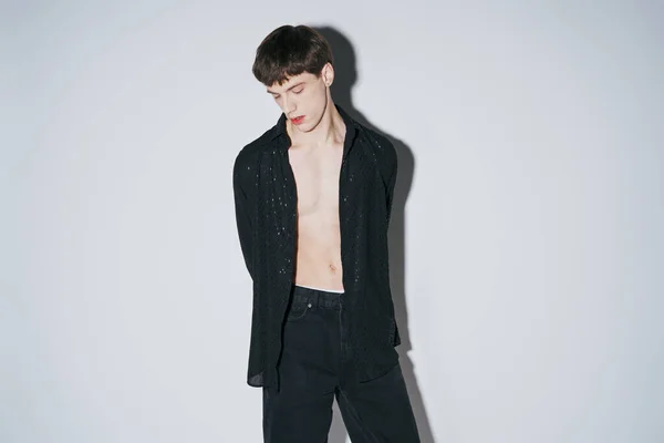 Confiant jeune homme en noir brillant chemise ouverte posant avec les mains derrière le dos sur fond gris — Photo de stock