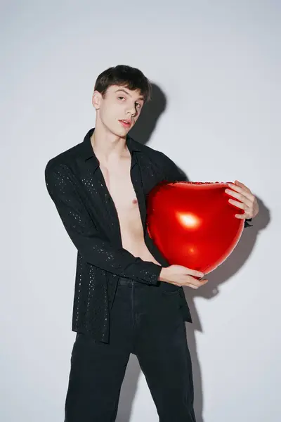 Joven con estilo en camisa abierta brillante negro sosteniendo globo rojo sobre fondo gris, día de San Valentín - foto de stock