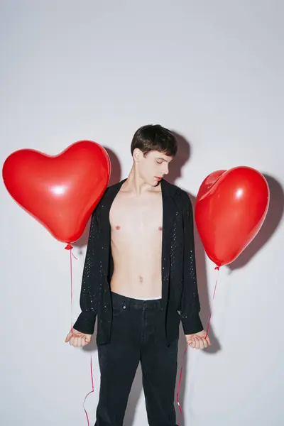 Jeune homme en chemise ouverte brillante noire tenant des ballons rouges sur fond gris, Saint Valentin — Photo de stock