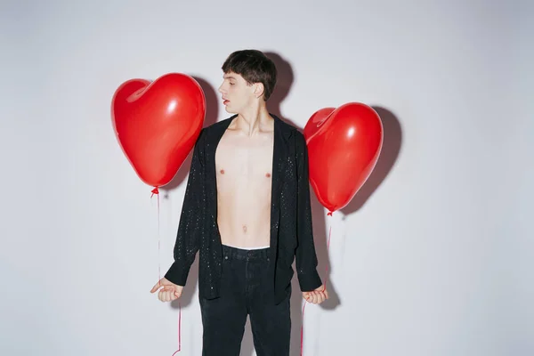 Romántico joven en camisa abierta brillante negro sosteniendo globos rojos sobre fondo gris, día de San Valentín - foto de stock