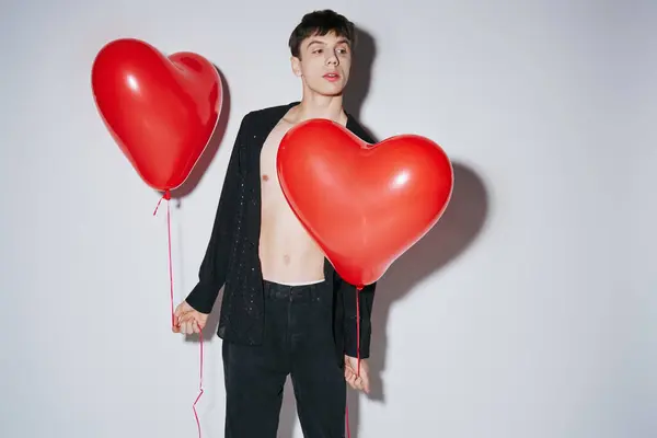 Romántico joven con camisa negra abierta sosteniendo globos rojos sobre fondo gris, San Valentín - foto de stock