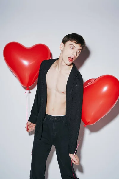 Young man in black shirt posing with open mouth and holding red balloons on grey background — Stock Photo