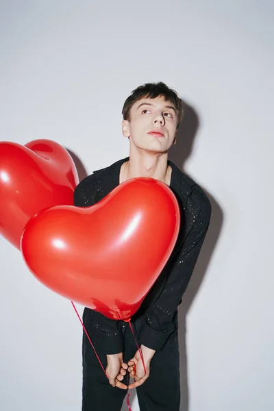 Young man in black shirt holding red heart shaped balloons on grey background, Valentines day — Stock Photo