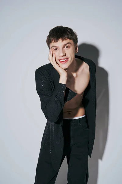 Fröhlicher junger Mann in gepunktetem schwarzem Hemd und Jeans posiert mit händennaher Wange auf grauem Hintergrund — Stockfoto