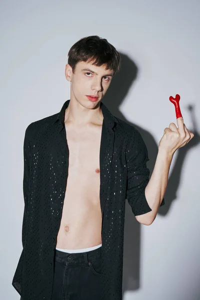 Provocative young man in open black shirt showing middle finger with red balloon on grey background — Stock Photo