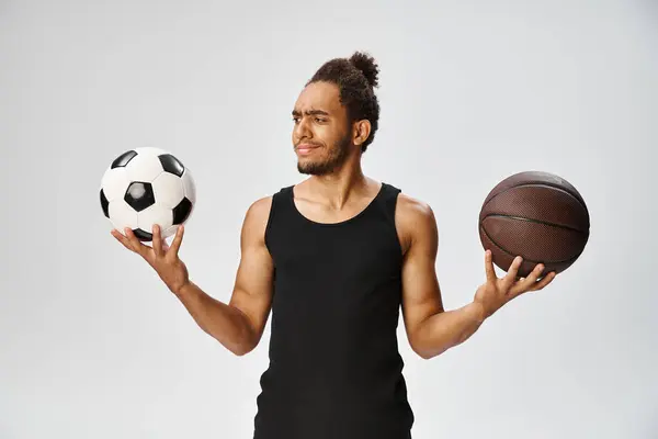 Confused athletic african american man in sportwear holding football and basketball on gray backdrop — Stock Photo