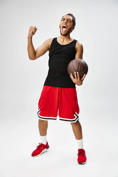 Emotional good looking african american sportsman posing with basketball on gray background — Stock Photo