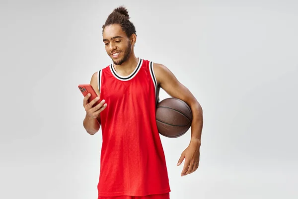 Gioioso sportivo afro-americano in possesso di basket e guardando il suo smartphone, scommesse online — Foto stock