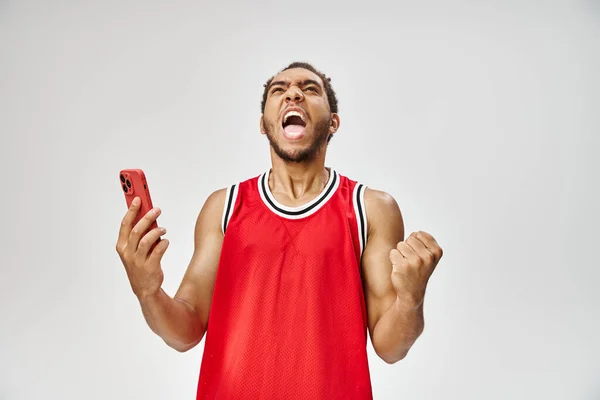 Emotional handsome african american man holding smartphone on gray backdrop, online betting — Stock Photo