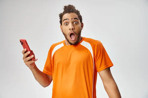 Shocked african american man in sportwear holding phone and looking at camera, online betting — Stock Photo