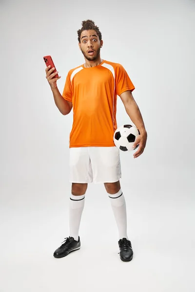 Shocked african american man holding phone and football and looking at camera, online betting — Stock Photo