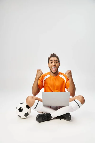 Fröhlicher afrikanisch-amerikanischer Sportler neben Fußball mit Laptop vor der Kamera, Online-Wetten — Stockfoto