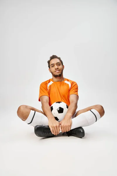 Cheerful african american man in cozy sportwear sitting on floor with football and looking at camera — Stock Photo