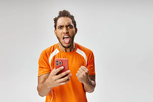 Sorprendido hombre afroamericano en uniforme deportivo con teléfono inteligente mirando a la cámara, apuestas en línea - foto de stock