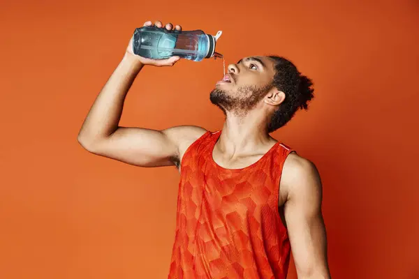 Добре виглядає спортивний афроамериканський чоловік п'є прісну воду, позуючи на помаранчевому фоні — стокове фото