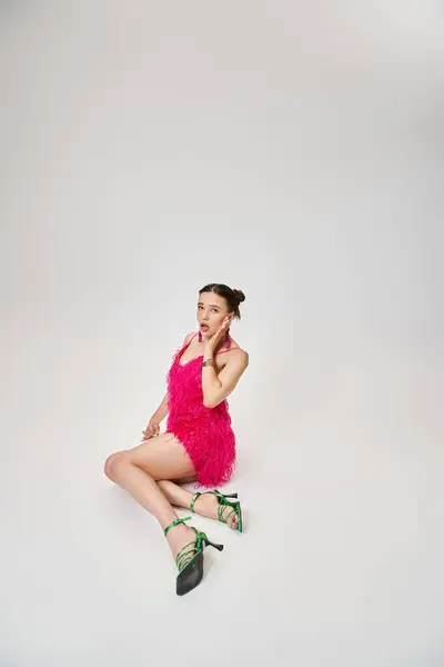 Chica juguetona en vestido rosa de moda y zapatos verdes tocando su mejilla y sentado sobre fondo gris - foto de stock