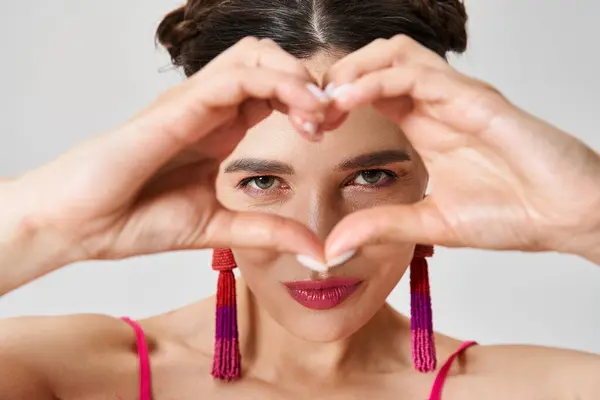 Joyful brunette woman wearing pink feather dress showing a hand heart and looking through it — Stock Photo