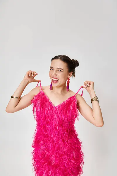 Hübsche lachende Frau im rosafarbenen Kleid, die in die Kamera schaut und Kleiderträger auf grauem Hintergrund hält — Stockfoto