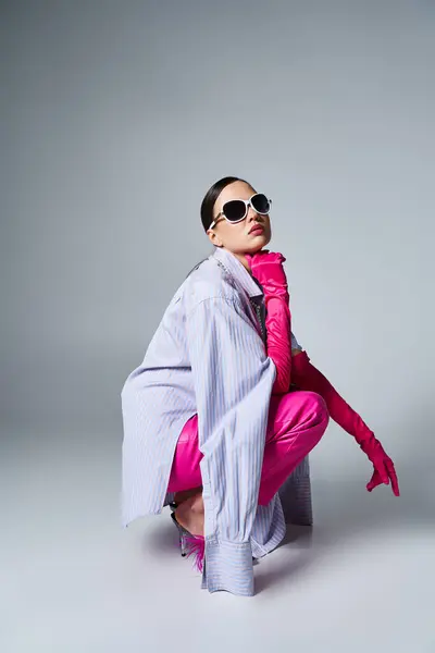 Selbstbewusste Brünette in pinkfarbener Lederhose und schicker Sonnenbrille sitzt elegant auf grauem Hintergrund — Stockfoto