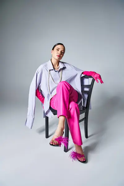Mujer morena bonita con guantes de color rosa y pantalones elegantes, sentado en la silla y mirando confiado - foto de stock