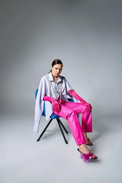 Chic chica morena en elegante traje rosa tocando sus piernas, sentado en la silla sobre fondo gris - foto de stock