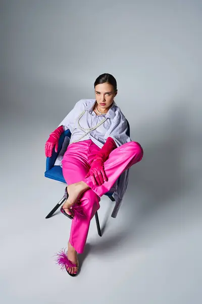 Elegante chica morena en traje rosa de moda sentada en la silla y sosteniendo su pierna, mirando a la cámara - foto de stock