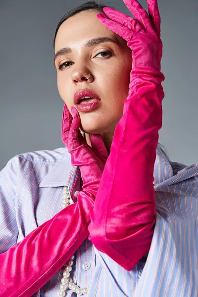 Брюнетка с пирсингом в стильном наряде и розовых перчатках, касается ее лица, глядя в камеру — стоковое фото
