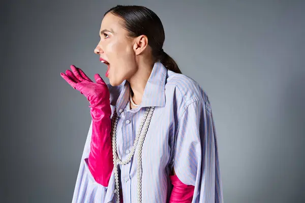 Femme moderne avec des gants roses, rit joyeusement avec des cris tout en faisant un geste sur fond gris — Photo de stock