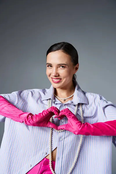 Smiling brunette woman with pink trendy gloves making hand heart isolated on grey background — Stock Photo