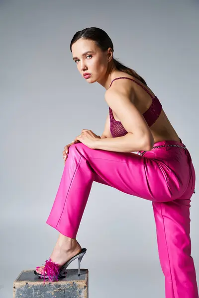 Stylish brunette in trendy red top, pink pants and feather shoes, touching her leg on grey backdrop — Stock Photo