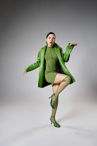 Brune fashion-forward en mini robe verte, veste en cuir, chaussettes à genoux dansant sur fond gris — Photo de stock