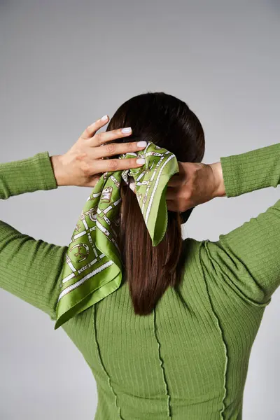 Back view of young brunette woman in total green outfit tying her head scarf on grey background — Stock Photo