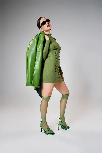 Femme sexy et élégante portant une tenue verte et des lunettes de soleil, tenant une veste en cuir sur son épaule — Photo de stock