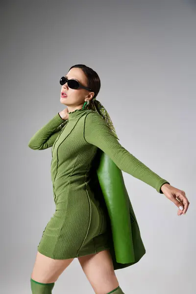 Chica morena en traje verde y gafas de sol, sosteniendo chaqueta de cuero en su hombro, dando un paso - foto de stock