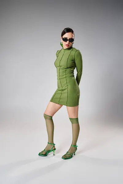 Jolie femme en mini robe verte et chaussettes de genou, portant des lunettes noires sur fond gris — Photo de stock