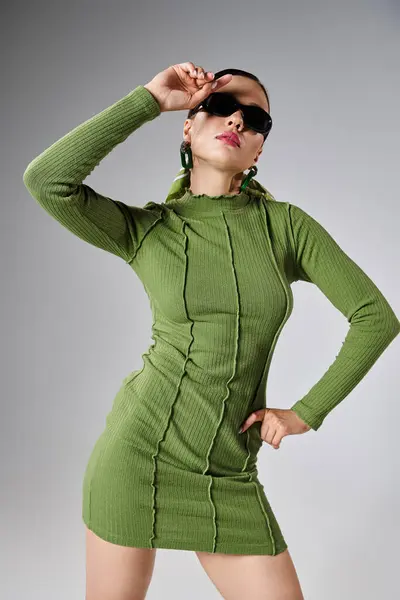 Morena extravagante em mini vestido verde usando óculos de sol e tocando sua testa em fundo cinza — Fotografia de Stock