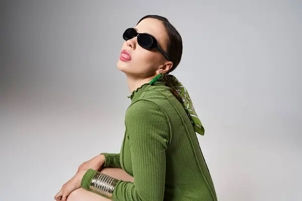 Vista lateral da senhora em roupa verde total usando óculos de sol sentados no fundo cinza mostrando a língua — Fotografia de Stock