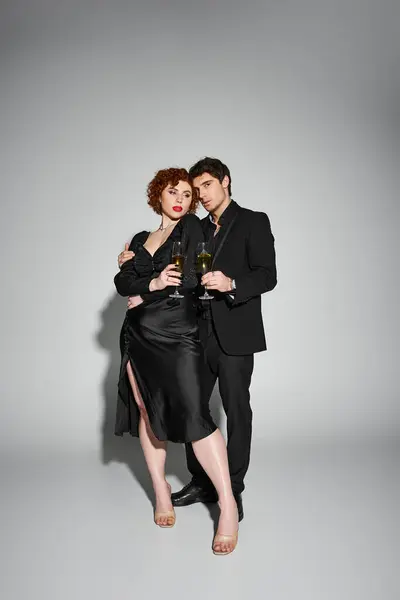 Schöne Frau in schwarzem Kleid umarmt ihren hübschen Freund vor grauem Hintergrund, Champagner in den Händen — Stockfoto