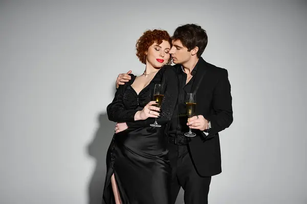 Beautiful woman in black dress hugging her handsome boyfriend on gray backdrop, champagne in hands — Stock Photo