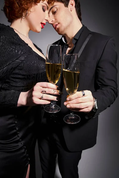 Verführerische Frau in schwarzem Kleid umarmt ihren hübschen Freund vor grauem Hintergrund, Champagner in den Händen — Stockfoto