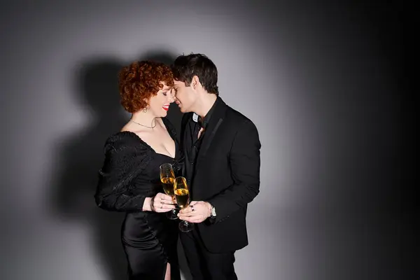 Atractiva mujer en vestido negro abrazando a su novio guapo en fondo gris, champán en las manos - foto de stock
