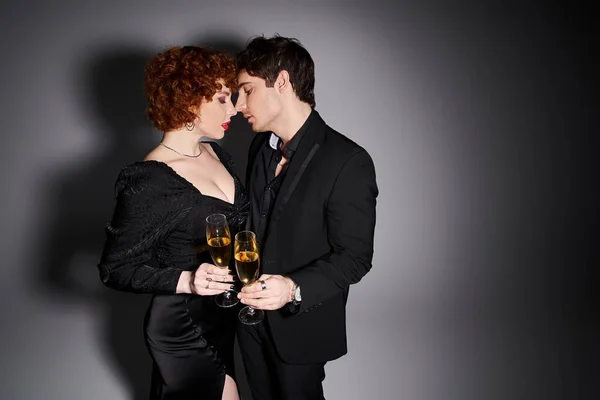 Attraktive Frau in schwarzem Kleid umarmt ihren hübschen Freund vor grauem Hintergrund, Champagner in den Händen — Stockfoto