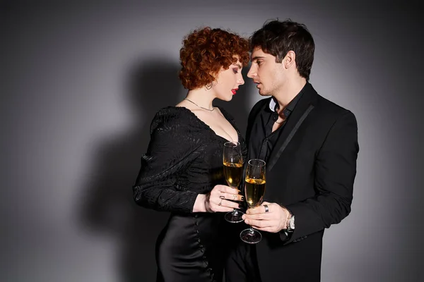 Attraktive Frau in schwarzem Kleid umarmt ihren hübschen Freund vor grauem Hintergrund, Champagner in den Händen — Stockfoto