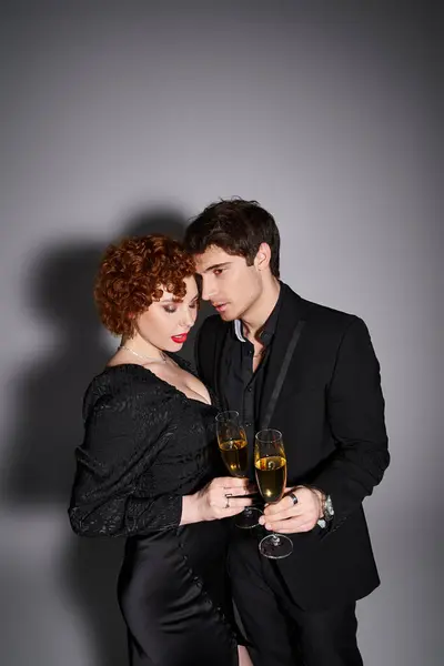 Charmante Frau in schwarzem Kleid umarmt ihren hübschen Freund vor grauem Hintergrund, Champagner in den Händen — Stockfoto