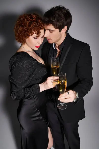 Charmante Frau in schwarzem Kleid umarmt ihren hübschen Freund vor grauem Hintergrund, Champagner in den Händen — Stockfoto